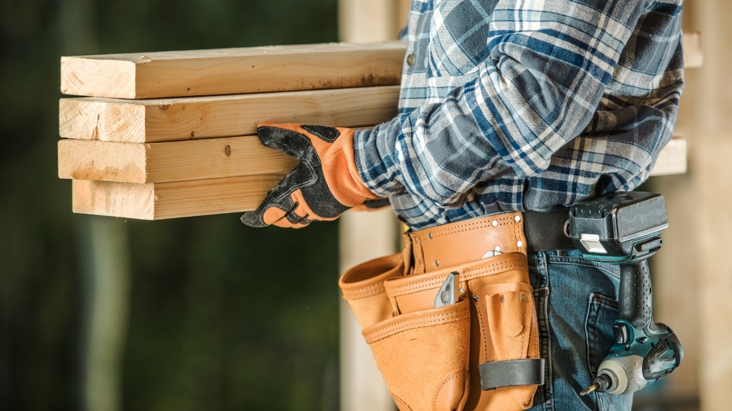 A man wearing gloves, a flannel shirt, and a utility belt carries lumber.