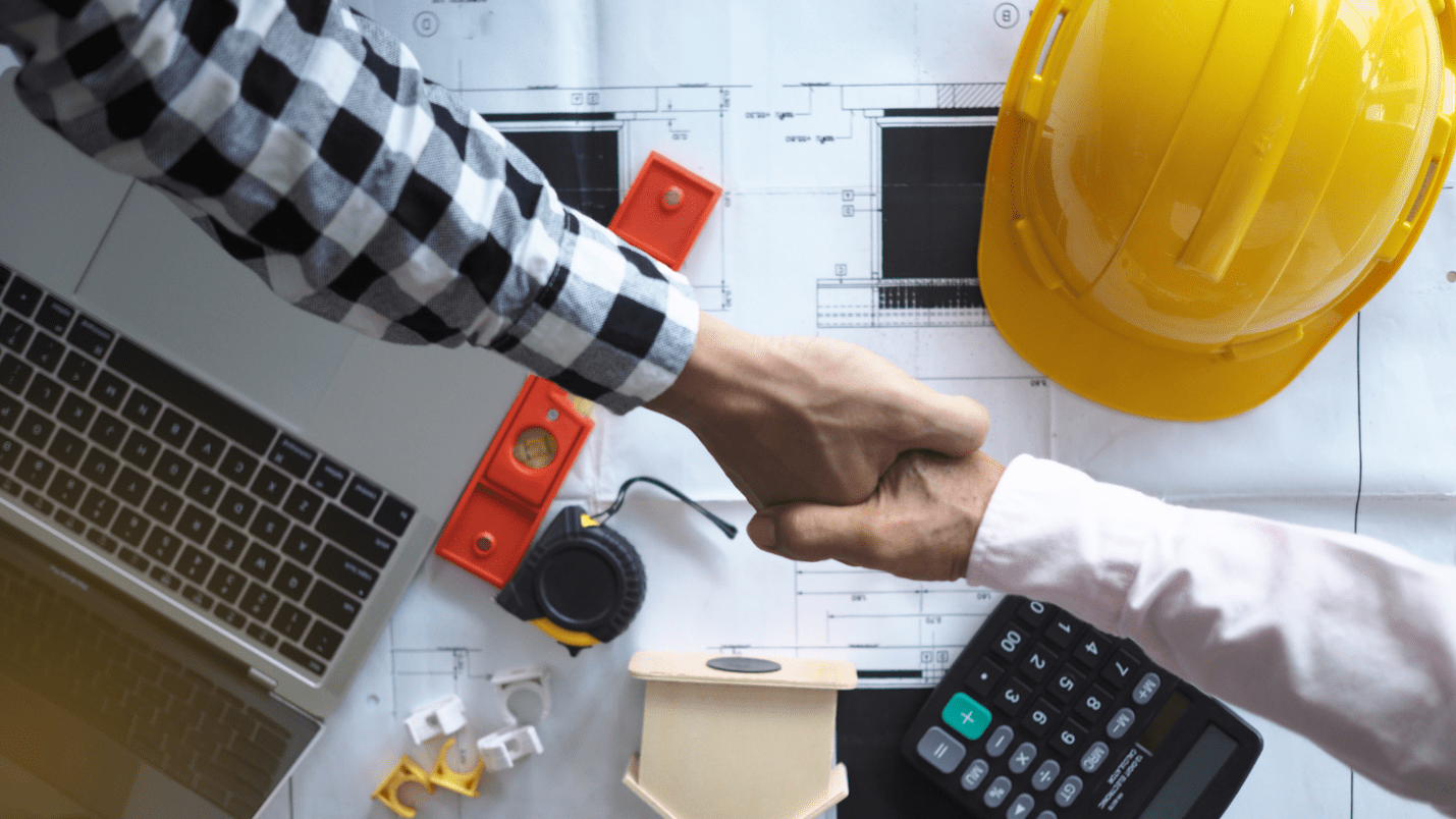 Two people shake hands, below the hands are blueprints and a hardhat.