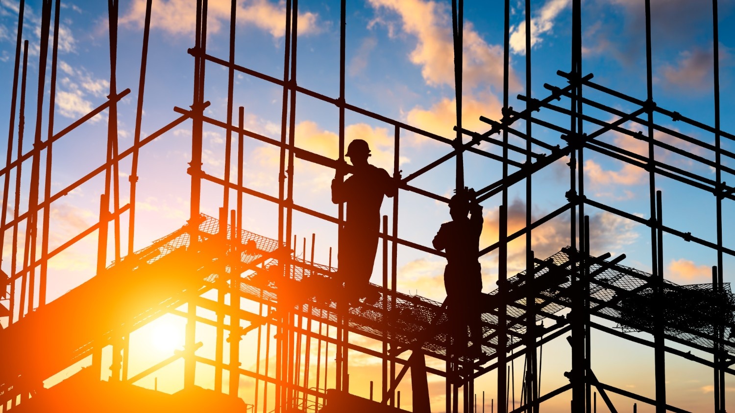 Two construction workers stand on scaffolding at sunset.